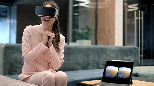 A blonde caucasian excited woman exploring virtual reality using VR glasses in an office. Tablet on