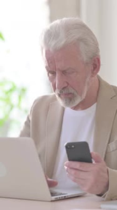Old Man Browsing Internet on Smartphone while using Laptop in Office, Vertical Video