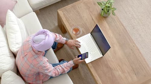 Utilisant un smartphone et un ordinateur portable, homme travaillant à domicile sur un canapé