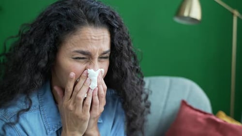 Close up portrait of young woman suffers from allergies sneezes with a runny nose sitting on sofa