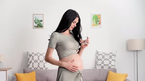Schöne schwangere Frau streichelt ihren Bauch mit der Hand und trinkt gesundes sauberes Wasser im