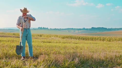 Happy Young Farmer in Hat Rejoicing and Looking at the Camera While Posing with a Shovel in a Field