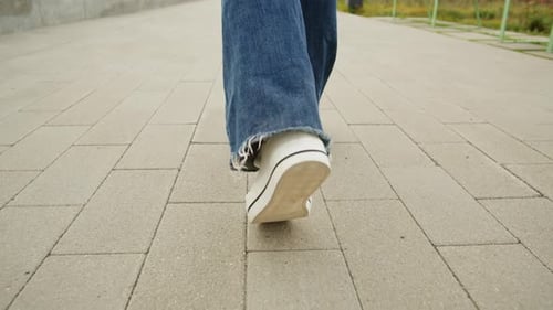 Young Woman Wearing Blue Jeans and White Sneakers Walking on Asphalt Road at Sunny Day Back View