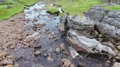 Ein kleiner Fluss fließt durch eine felsige Landschaft mit grünem Gras und moosbewachsenen Steinen