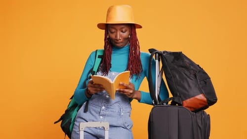 Woman Reading with Luggage on Orange Background