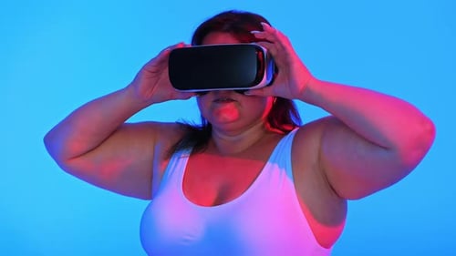 Woman with overweight in a tracksuit putting on VR headset in a studio with blue background and red