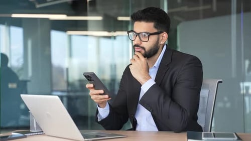 Thoughtful businessman is using mobile phone sitting at workplace in business office.