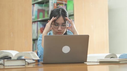 Tired Student Massaging Temples in Library with Laptop