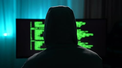 Rear View of Unrecognizable Male Hacker Wearing Sweatshirt with Hood Typing on Keyboard Looking to