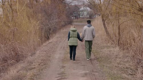 A Young Couple Walks Hand in Hand Along a Dirt Path on a Cloudy Spring Day They are Dressed in