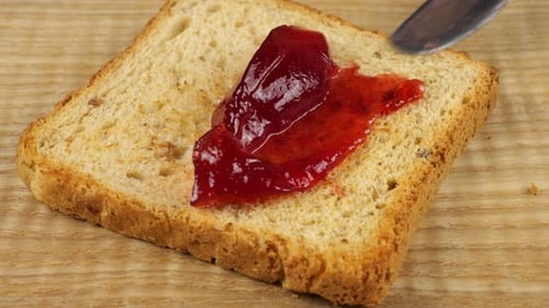 Geléia de groselha vermelha é espalhada com uma faca sobre uma fatia de pão torrado multigrão em uma tábua de madeira