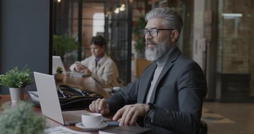Experienced Businessperson Using Laptop Computer Then Answering Mobile Phone Call Working in Cafe