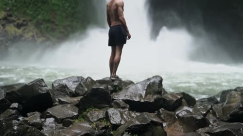 Man looks around as jungle waterfall crashes down behind him, slow motion