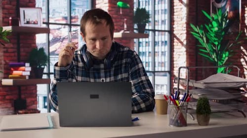 Stressed Man in Home Office Scratching Head with Pen Feeling Overworked