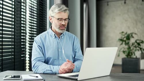 Mature gray haired bearded businessman talking on a video call using a laptop at a workplace