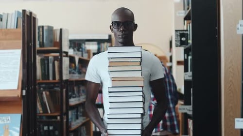 Black Male Student Stands with a Stack of Books in Library Against the Background of Students