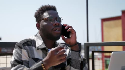 Portrait of Black Man Talking By Mobile Phone Working Remotely with Laptop Computer in Cafe
