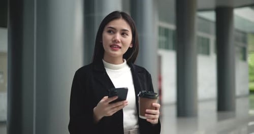 Young elegant Asian busy business woman in a suit holding mobile phone and a cup of coffee