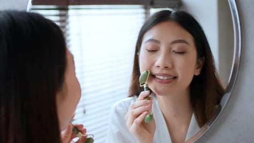 Woman Using Jade Facial Roller in Bathroom Mirror