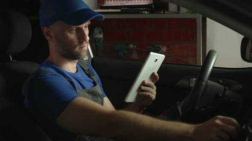 Ein Mechaniker sitzt nachts mit einem Tablet-Computer auf dem Fahrersitz eines Autos in einer Garage