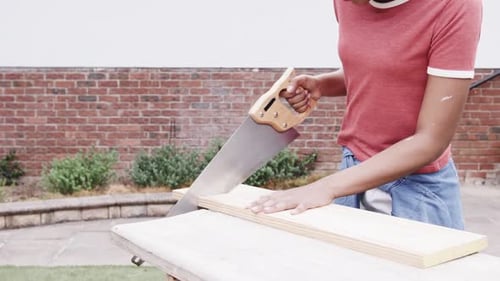 African American woman steadying wood, donning goggles, sawing with saw for DIY, patio, copy space