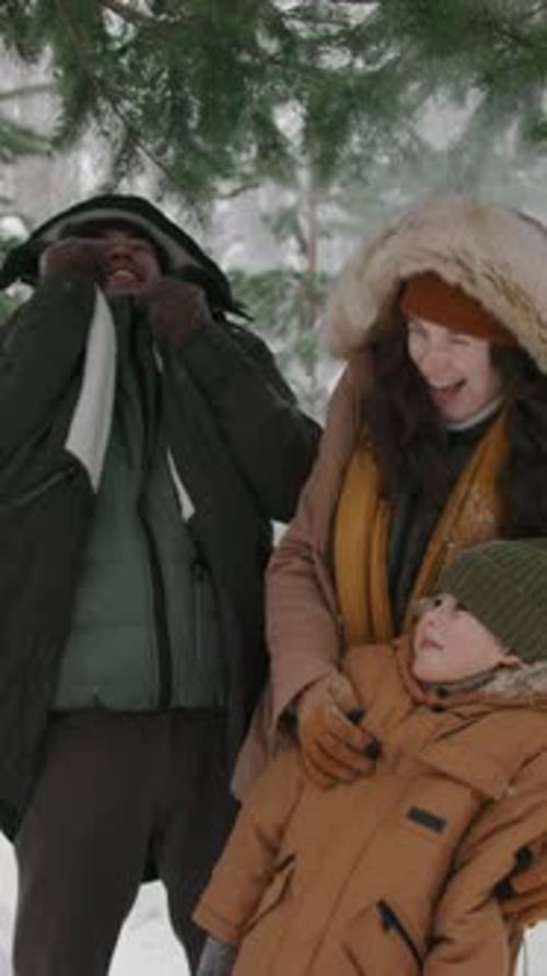 Young Family Standing under Falling Snow in Pine Forest and Laughing