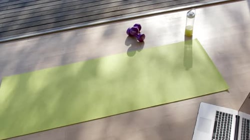 High angle view of yoga mat, laptop, bottle with juice and weights on floor at home, slow motion