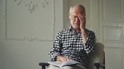 Senior Man Reading Book has Headache Indoors