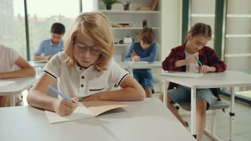 Redhead School Boy Wearing Glasses Sitting on the Desk Writing the Dictation