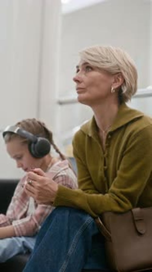 Stressed Woman Sitting next to Daughter in Headphones while Waiting in Hall