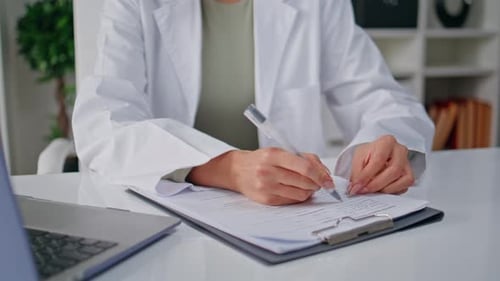 Physician Hands Writing Records Sitting Clinic Table with Laptop Closeup