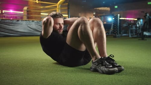 A Man is Energetically Performing Crunches on Artificial Turf Next to a Boxing Ring Inside a Gym