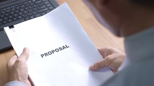 Man Reads Proposal Document at Desk with Laptop