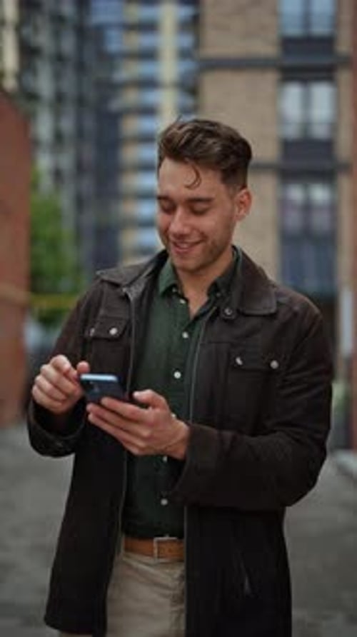Vertical Portrait of Happy Young Man Using Mobile Phone While Walking Along City Street Smiling