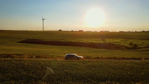 SUV Driving Along Dirt Road Surrounded By Green Fields Under Warm Sunlight Leaving Trail of Dust