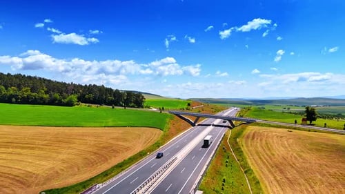 Sunny road through green fields. Trucks and cars travel along a wide highway surrounded