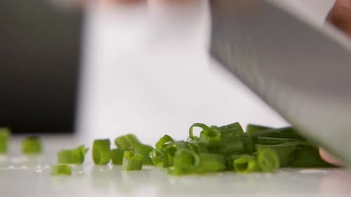 Close-up of green onions being finely chopped on a white cutting board, knife in motion