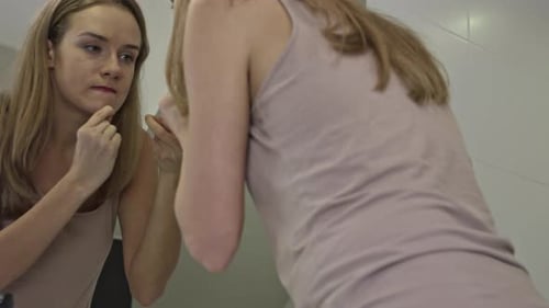 Young woman squeezes pimples on her chin while standing in the bathroom in front of the mirror.