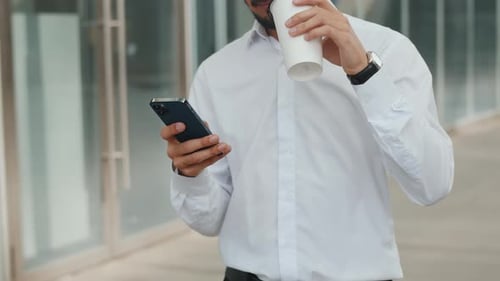 Young Businessman in a White Shirt Walks Near the Business Center with a Smartphone and Drinking