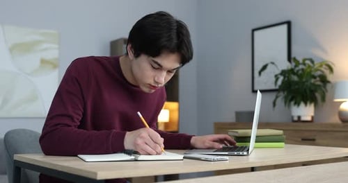 Young student studying with laptop and writing in notebook at wooden table indoors
