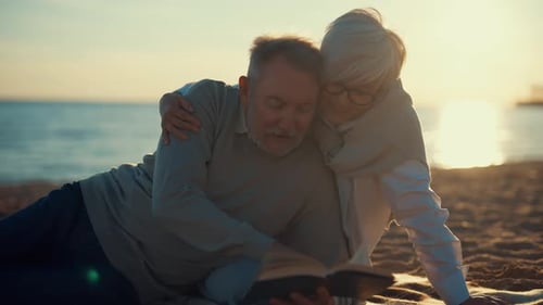Senior Gray Haired Man Woman Reading Book Together Sitting on Sandy Sea Beach at Sunset in Evening
