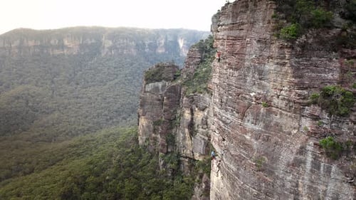 Rock climbers scaling a huge steep mountain above a green valley. He slowly struggles and makes his