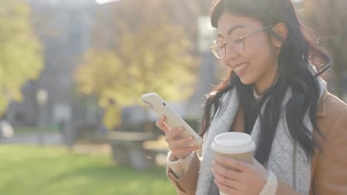 Smiled Asian Woman Walking in the Park with Cup of Coffee Using Smartphone