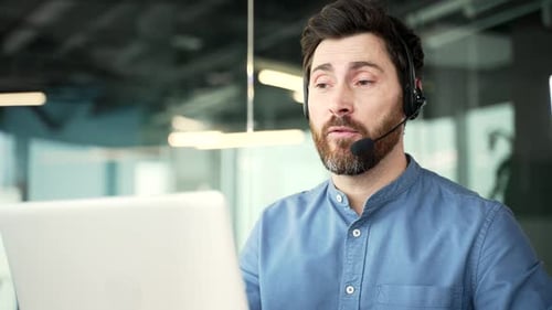 Confident businessman in headset talking on a video call using laptop sitting at workplace in office