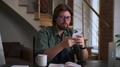 Young Man Talking and Using Smartphone While Sitting at Table in Home Office Spbas