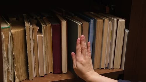Hand Touches Spine of Books on Wooden Shelf