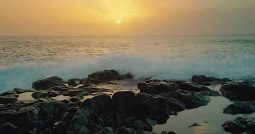 Ocean waves crash on shore covering the setting sun on the horizon. Close-up rocky beach stormy sea