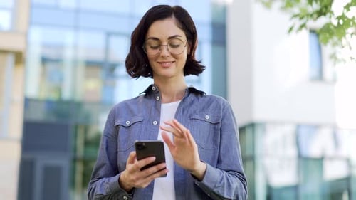 Smiling female student using smartphone standing in campus space near university building. Brunette