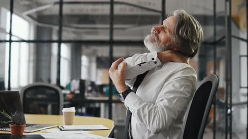 Tired mature businessman with grey hair and beard in formal clothes is in the office uses laptop