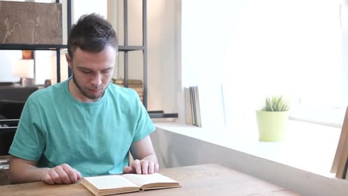 Young Man Reads Book at Table Indoors